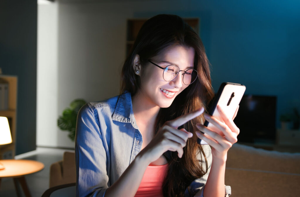 A young woman wearing a pair of blue light glasses while using her phone in a dimly lit room.