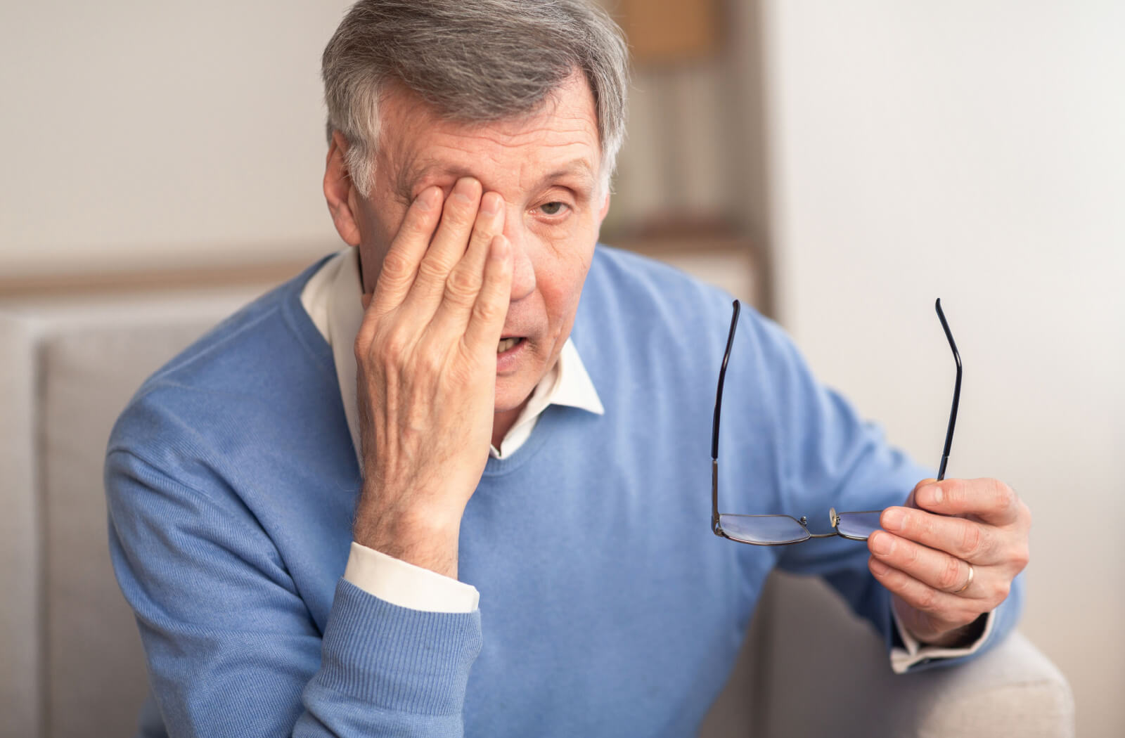 A senior man sitting on a couch and holding his glasses in his left hand as he rubs his right eye with his other hand.