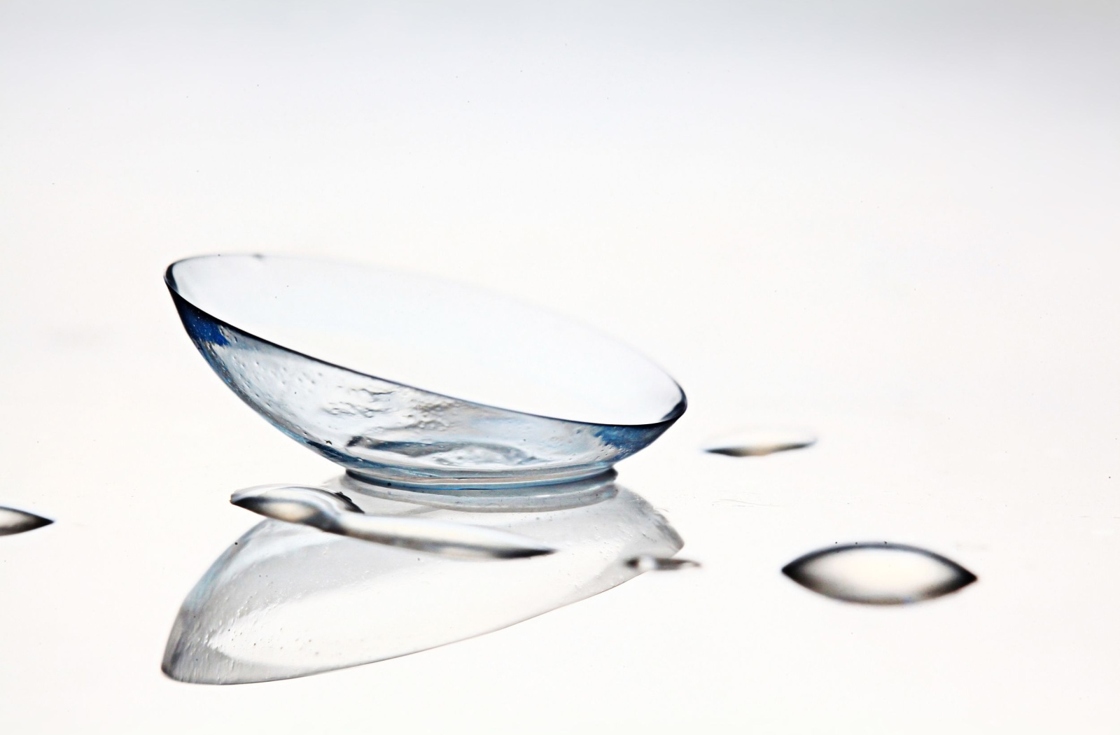 A contact lens sitting on a white surface with drops of contact lens solution surrounding it.