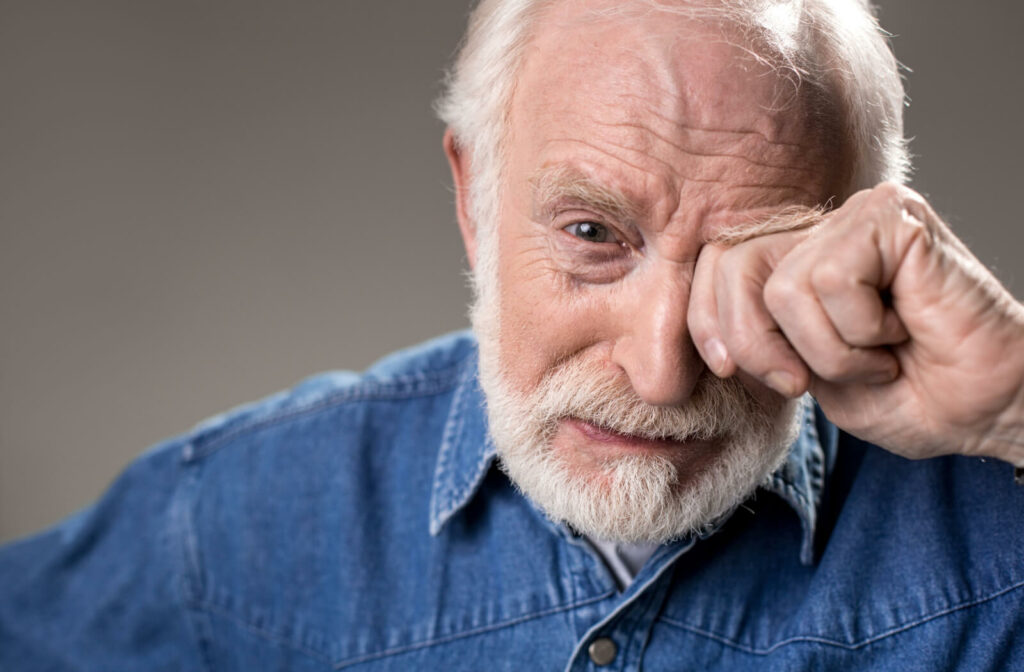 A senior adult man rubbing his left eye with his left hand.