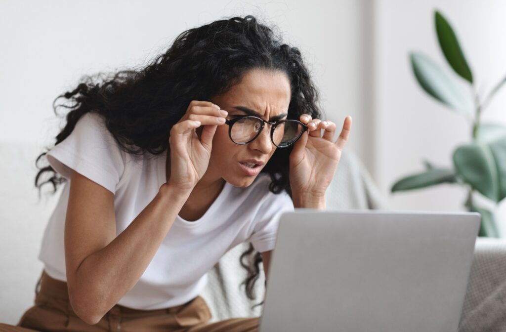 A woman is squinting by her computer with her glasses due to eyesight declining.